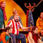 A man in a red and white striped football shirt is sitting with his arms and hands above his knees, as if waiting or listening for something. A group of people sit around him, listening to what he has to say. Behind them is a large yellow sunset.