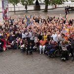 A large group of people sit and stand outside with arms in the air.