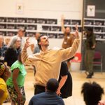 A man in a beige hoodie dances with arms out.