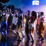 A dance company performing on the open air River Stage, Seated audience members, trees, sky and an illuminated NT logo beyond.