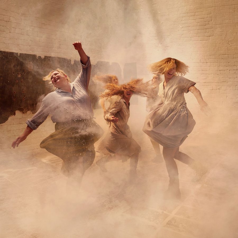 A group of women dancing exuberantly, kicking up dust around them, smiling, wearing light cotton dresses and skirts