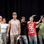 The actor John Partridge surrounded by six members of the company standing in a V formation with one arm raised.