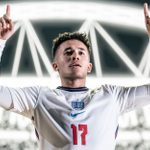 A man stands, both hands raised, index fingers pointing up. He wears a white England shirt with the number 17. A football stadium is projected behind him.