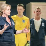 A woman in an England tracksuit stands, arms outstretched, talking. A man in a yellow goalkeepers kit stands behind her, looking at her. Another man in an England tracksuit and white shirt stands behind her, looking away.