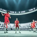 A group of men are wearing red England football kit. One leaps as if to head a ball and the others are standing round him, watching and anticipating the result of the header. The image of a football stadium and the words TUN 0 - 0 ENG, repeated, are projected behind them.