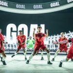 A group of men, most wearing red England football kit and one in a green goalie uniform, are in various stages of celebration after a goal. The image of a football stadium and the word GOAL is projected behind them.