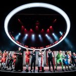 A number of people wearing a mix of football kits, tracksuits, suits, ecclesiastical clothing, flags, are standing in a semi-circle, most looking excited. Above them is a white circle and in the inside is projected CRO 2 - 1 ENG, repeated, in red on black.