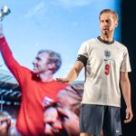 A man stands, hands on hips, he wears a white England shirt with the number 9, his mouth is open, talking, and he stares straight ahead. Behind him is a film still of Bobby Moore holding the World Cup aloft.