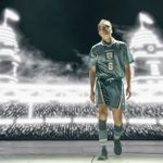 A man walks towards us, wearing a grey England football kit with teh number 6 on it. Behind him is projected a football stadium with twin towers.