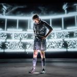 A man stands with his hands on his hips, looking down at the ground. He wears a blue football strip from 1996. He is up-lit, behind him is a projection of a stadium.