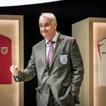 A man stands in a grey suit with the white England emblem of the three lions his arm is extended with a clenched fist. He has grey hair. Behind him is a white England strip shirt and a red England strip shirt.