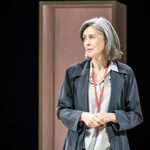 A woman stands, she has grey hair is a long bob. She is wearing a dark over coat with a grey shirt underneath, A red lanyard is around her neck. he hands are held together in front of her. She has a slight smile.