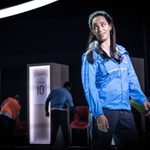 A woman with brown long hair which is half tied up stands. She wears a bright blue England windbreaker with the emblem on the chest. She is looking to the side, her hands hanging down.