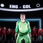 A man stands with his knees bent, he wears a green goalie uniform. His hands are ready to catch a ball, he is wearing goalie gloves, his face looks determined. He has short blonde hair. Behind him the rest of the players stand bent over focused, wearing the red England strip. above their heads shows a penalty score between ENG and COL