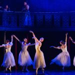 Four ballet dancers perform on a darkened stage, with musicians playing in a gallery above.