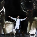 Mark Rylance, in shirtslevees and an operating apron, arms outstretched, surrounded by dancers.