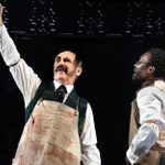 Two men wearing workers aprons over waistcoats and dress-shirts, both standing and smiling, and one raises an arm in celebration.