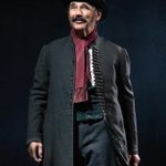 A man wearing an old fashioned coat a top hat and scarf, stands, smiling, holding a small suitcase.