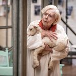 A woman holds a stuffed animal lamb and looks off with a concerned look.