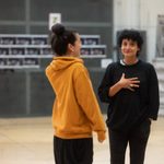 Two women stand talking. One woman wears a bright yellow hoodie and the other wears a dark sweater.