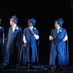 Four actors on stage dressed in striped blue period costumes with top hats, engaged in a lively conversation, against a stark black backdrop.