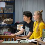 A man and a woman sat at the edge of the room, holding scripts in their laps. They are looking forward, watching rehearsals.