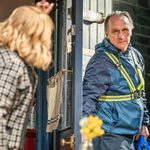 A man in a doorway, wearing a cycling jacket and high-vis webbing, looking into the room at a figure, is gesturing with a raised hand and pointing finger, talking to him.