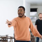 Fisayo Akinade, standing in a white space with blurred figures in the background, speaks and reaches out an arm.