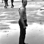 A white teenage boy in a t-shirt, sweater vest and black trousers looks at the camera. He stands in a puddle of water on a concrete playground. Other boys run around in the distance behind him. Black and white photograph.