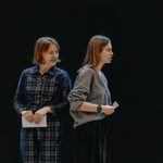 Two women holding slips of paper. The woman on the left has short brown hair and is wearing a blue plaid jumpsuit. The woman on the right has brown hair and is wearing a grey jumper. She has her back to the other woman and an upset expression.