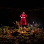 Actress in a striking red dress stands center stage, hands on hips, surrounded by an array of moorland flora, against a wooden backdrop, illuminated by spotlight.