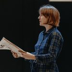 A woman with short brown hair, wearing a blue plaid jumpsuit and holding a newspaper. She is looking over the top of the newspaper with an upset expression.