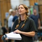 A woman with her hair in two braids and a pencil tucked behind her ear. She is turning the page of her script while acting with an upset expression.