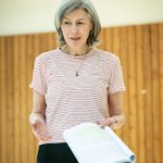 A woman with a grey long bob tucked behind her ears stands, she is looking down towards the right hand side. She is holding a script in her hand. She is wearing a red and white striped t-shirt and black trousers. she wears a simple necklace.