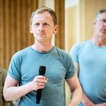 A man stands in a blue t-shirt, he is holding a microphone, he looks to the left, his face is straight. he has strawberry blonde hair. Behind him stands another man in a blue t-shirt who's face is blurred by the focus. Next to him stands a woman who is wearing a green shirt with a button polo neck, her dark hair is up in a ponytail. She is also obscured by the camera focus.