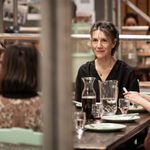 Harriet Walter sits at a table with a smile on her face. There is wine on the table and several other women sit around her.