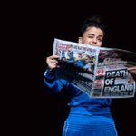 A young white woman, her brown hair tied up, dressed in a navy Adidas tracksuit and gold hoops, faces the audience while holding an opened newspaper with the title 'Death of England.