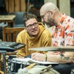 Two men sat next to a drum-kit and a sampling pad, having a conversation. One of the men is making notes into a book.