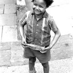 A young Black boy in a striped shirt, shorts and socks smiles at the camera. He holds an empty glass Cola bottle. Black and white photograph.