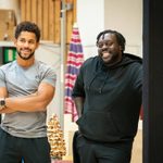 Two men stand next to each other they are smiling. The man on the left is wearing a gret tshirt and has a light black beard. He has his arms folded. The man next to him is wearing a black hoodie he also has a light black beard.