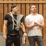 Two men in rehearsals. The man on the left is wearing black casual clothes and a black baseball cap on backwards. He has ropes tied to grey fabric draped over his shoulders and is holding a script. The man on the right is wearing a white t-shirt and is rubbing his hands together.