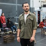 A young man with dark hair and a short beard, wearing a green shirt with the sleeves rolled up to the elbows and a red pencil tucked into the breast pocket. Behind him, other members of the cast are stood and sat at the edge of the room, watching.