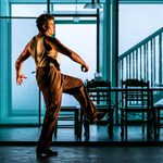 A man in a beige top and brown trousers dances in a house.