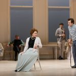 Janie Dee sits on a chair in the middle of a rehearsal room. She is wearing a light beige cardigan, a black top, a long light green skirt and a white pearl necklace. Johnny Flynn stands beside her.