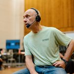 A man wearing a light green T-shirt and blue jeans sits on a cardboard box. He also wears black headphones with a mic.