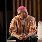 John Macmillan, hair dyed pink, wearing a striped fleece, sits holding a glass of wine, looking off to the right.