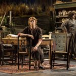 Justine Mitchell as Kate sits in a chair at a wooden table wearing a long brown dress. Siobhán McSweeney as Maggie stands in the background in a long grey floral cardigan.