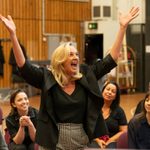 A woman holds her hand aloft in the air whilst looking up, mid song she has her mouth open in a smile. She wears a black blaze, black top and black and while gingham trousers. Around her are a group of women who are all sat on chairs.