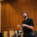 A man with dark brown hair and beard is gesturing mid-speech. He wears a black polo shirt and black trousers.