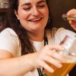 A smiling bartender with long dark hair pouring a pint of beer.